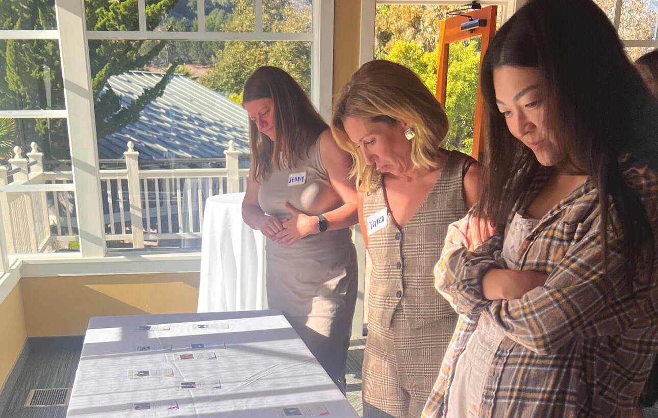 Three women standing by a sunlit window, reading workshop materials together