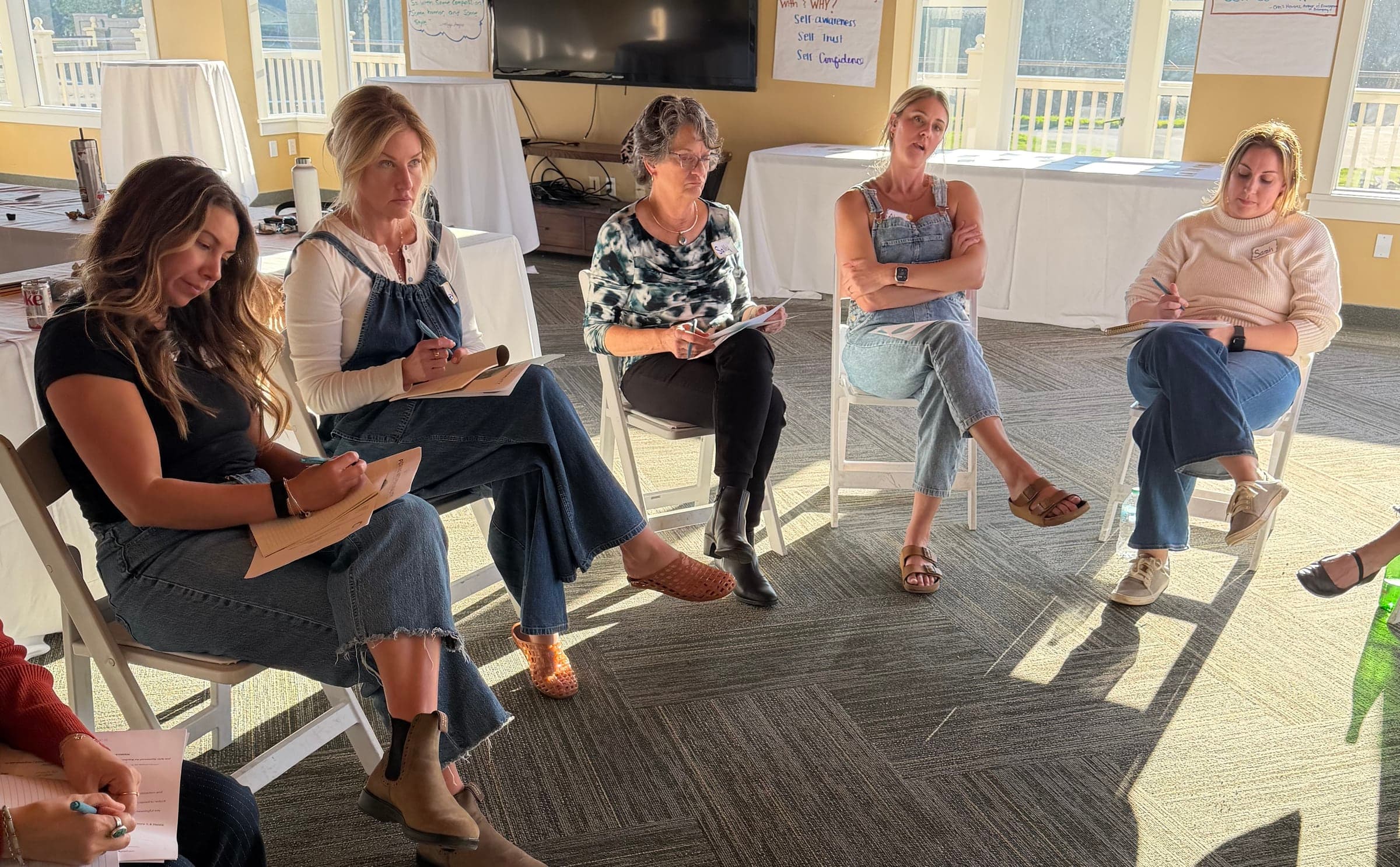 A participant reads her worksheet in the workshop circle, a hand-lettered chart of self-awareness, self trust and self confidence behind her