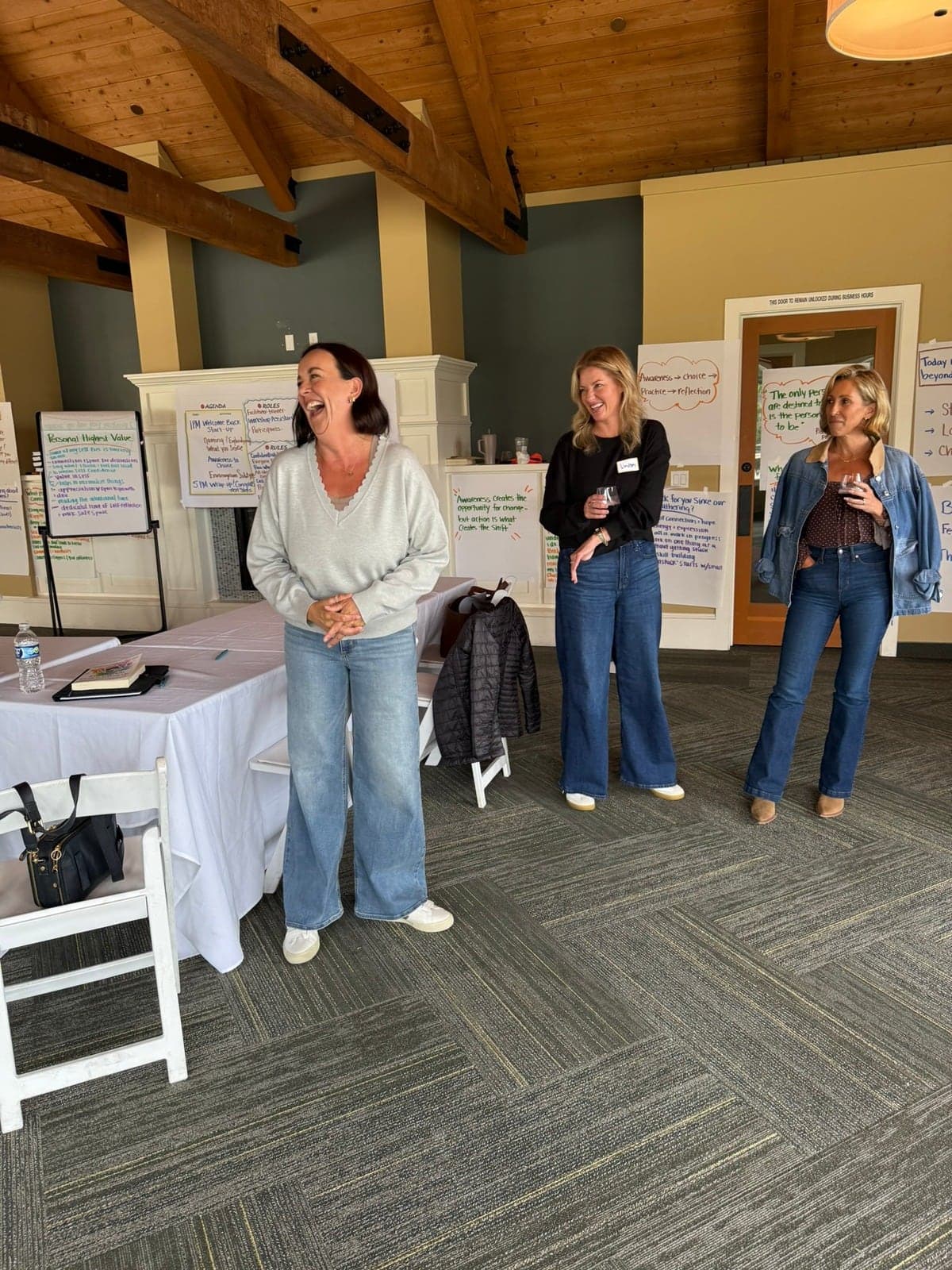 Katie laughing while facilitating a workshop, two participants with wine glasses behind her