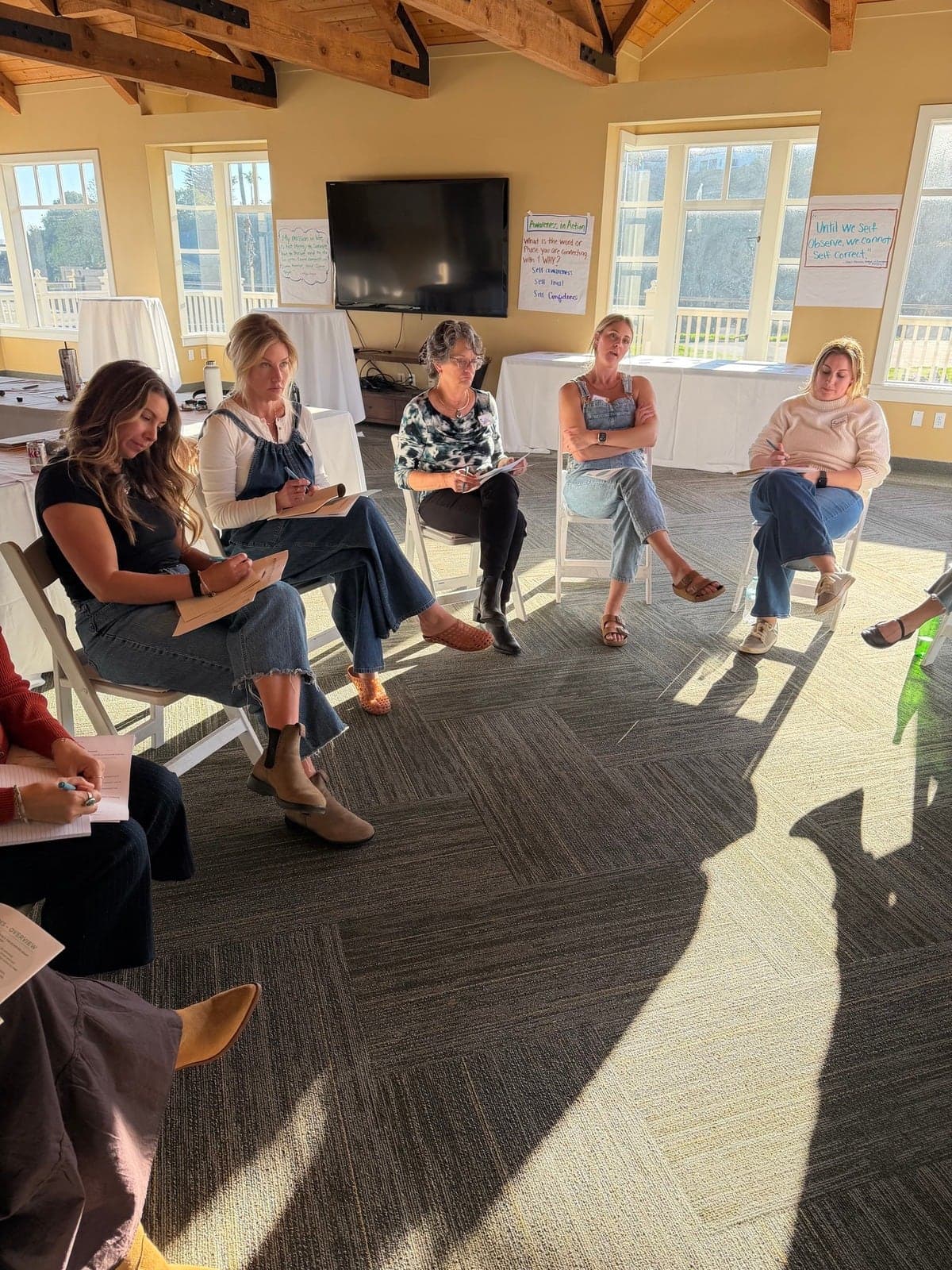 Five women seated in a circle in a bright workshop room, low sun through the windows and notebooks open